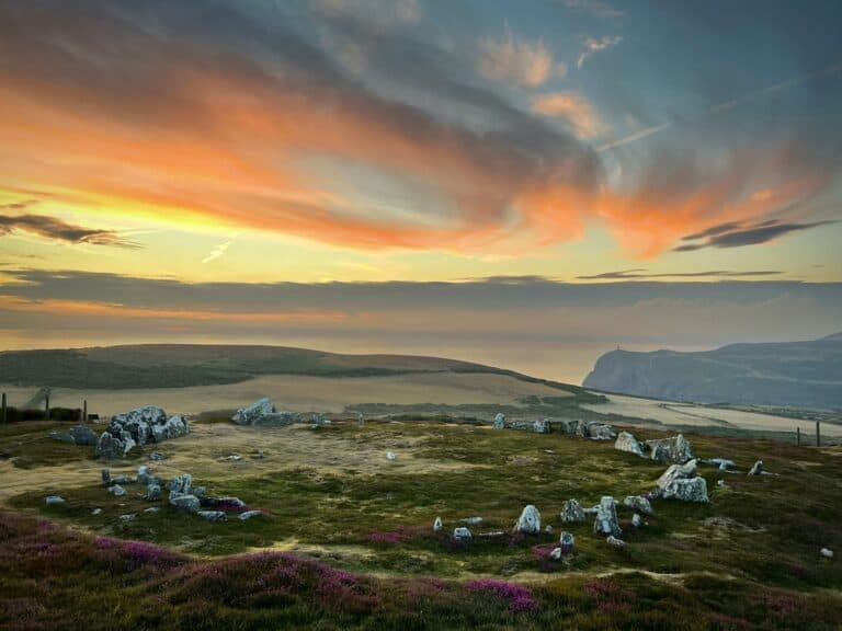 Stone circle on a hill with grass and purple heather at sunset, overlooking distant cliffs and a colorful sky.