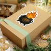 A sticker of a black cauldron with a spiral design, emitting stylized flames, on a wooden gift box.